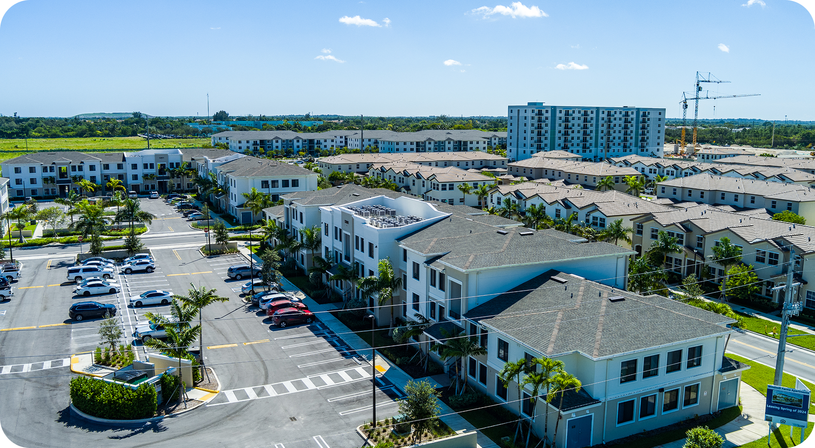 Aerial view of a shopping center parking lot with white storefronts, parked cars, and surrounding buildings.