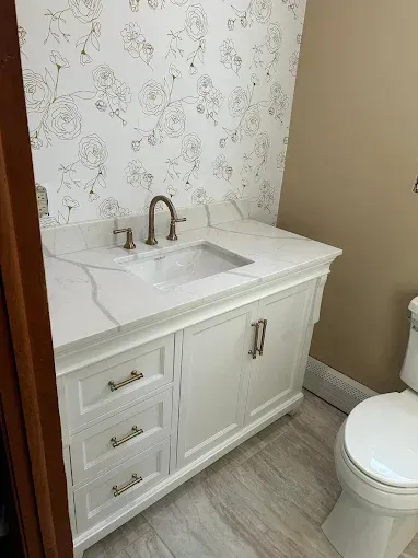 White bathroom vanity with marble countertop, bronze faucet, and toilet beside wallpapered wall.