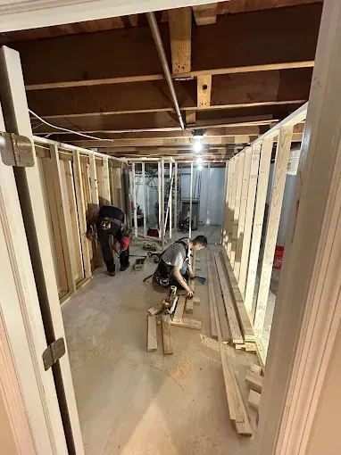 People framing interior walls in an unfinished basement, with lumber, tools, and exposed ceiling joists.