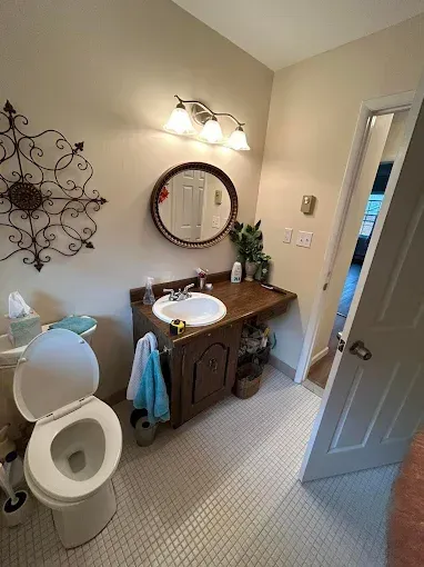 Small bathroom with toilet, wooden vanity, round mirror, wall sconce, and decorative wall art.