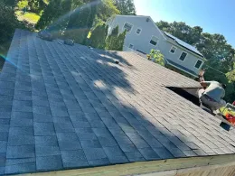 Worker installing shingles on a gray roof beside a white house on a sunny day