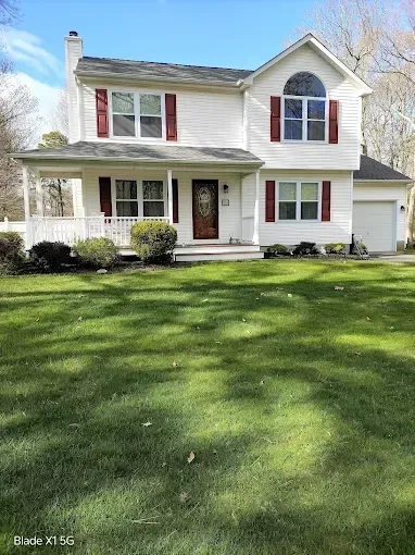 Two-story white house with red shutters, front porch, and a green lawn under a blue sky