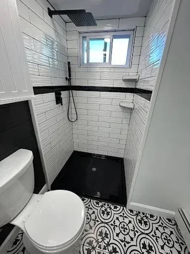 Small tiled bathroom with toilet, black shower tray, patterned floor, and window above the sink area