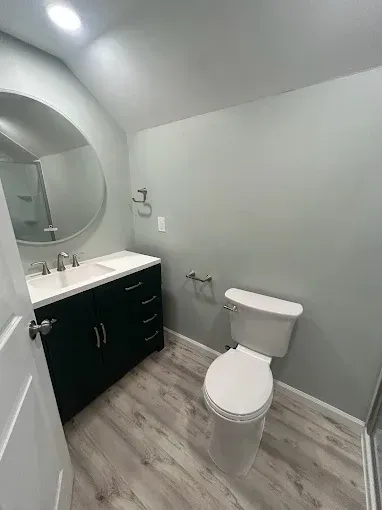 Small modern bathroom with white walls, gray floor, black vanity, round mirror, and toilet
