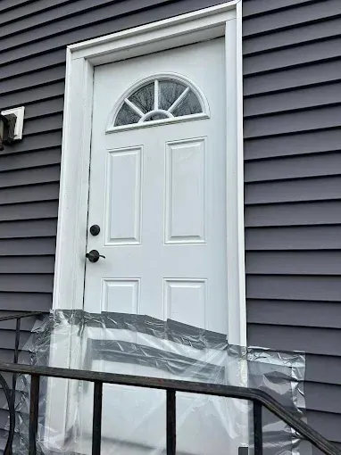 Front door with white trim and arched window, wrapped in clear protective plastic beside dark gray siding.