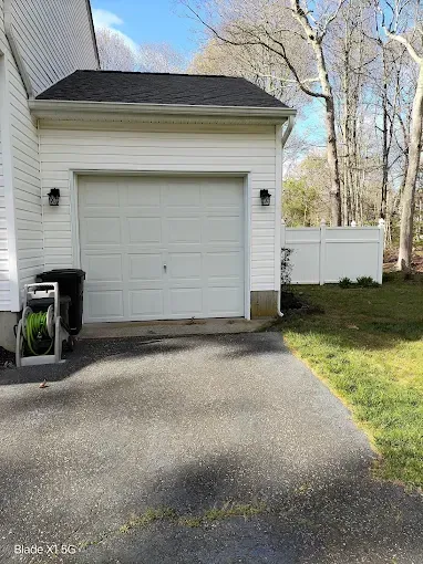 White garage with driveway, trash bins, and a grassy yard beside leafless trees.
