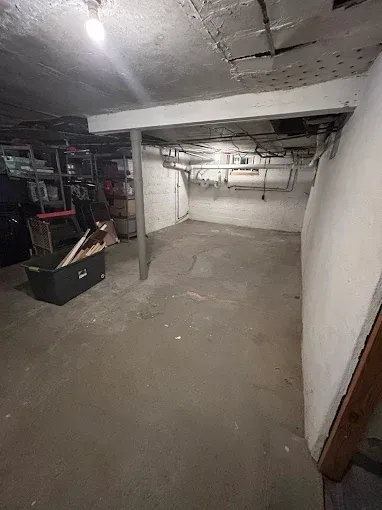 Empty basement with concrete floor, exposed pipes, shelves, and low ceiling under dim lighting.