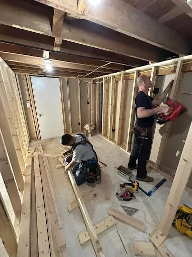 Two workers framing a basement wall, one kneeling with tools and one holding lumber in a partially finished room.