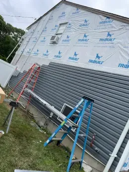 House under construction with house wrap, gray siding, and ladders leaning against the wall.