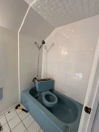 Small blue toilet in a compact tiled bathroom with a showerhead above.
