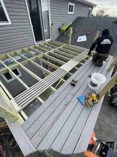 Worker building a rooftop deck frame beside a gray house, with tools and lumber on the deck.