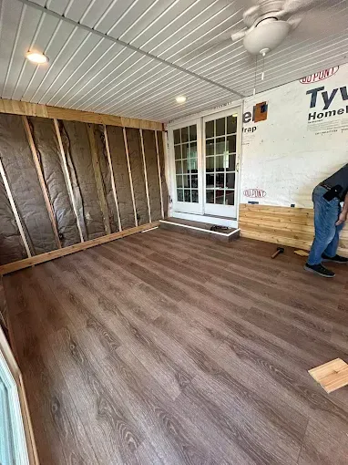 Empty room with wood-look flooring, white walls, windows, and a person working near the right wall.