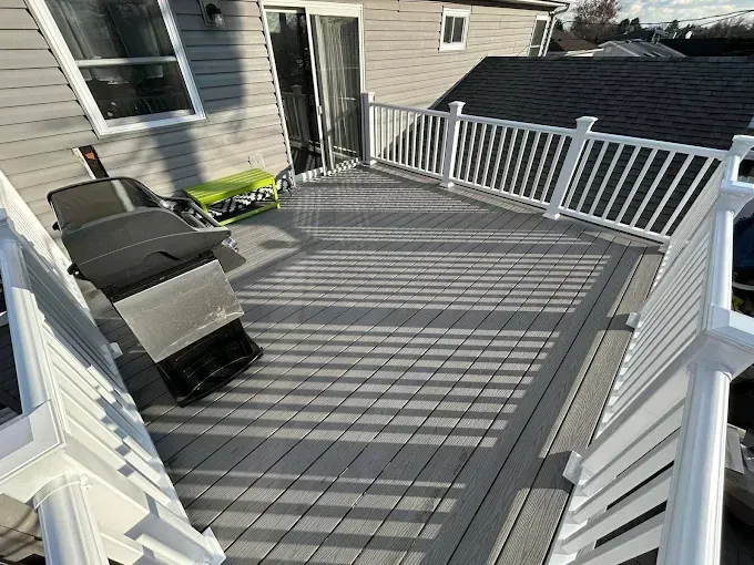 Gray deck with white railings and outdoor chairs beside a house in bright sunlight