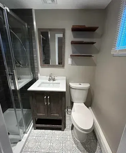 Compact bathroom with shower, vanity sink, toilet, wall shelves, and a small window