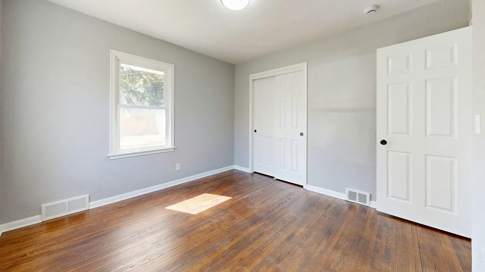 An empty bedroom with light gray walls, dark wood floors, a window on the left, and two white interior doors.