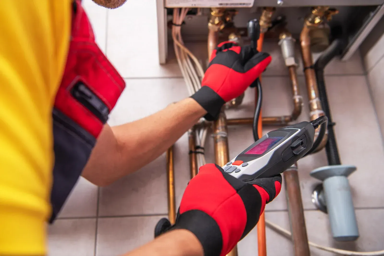 A worker in a yellow and red uniform uses a handheld diagnostic tool to inspect pipes inside a boiler unit.