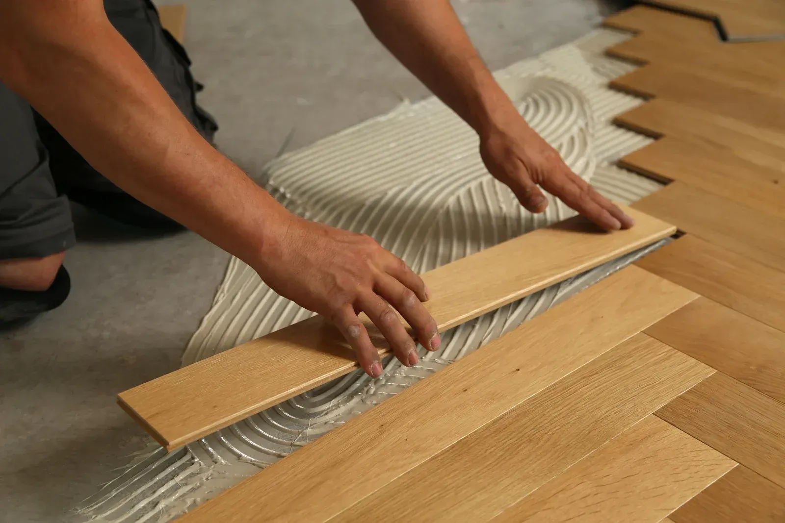 A person kneeling on the floor, carefully laying a wooden plank onto a bed of adhesive in a herringbone pattern.
