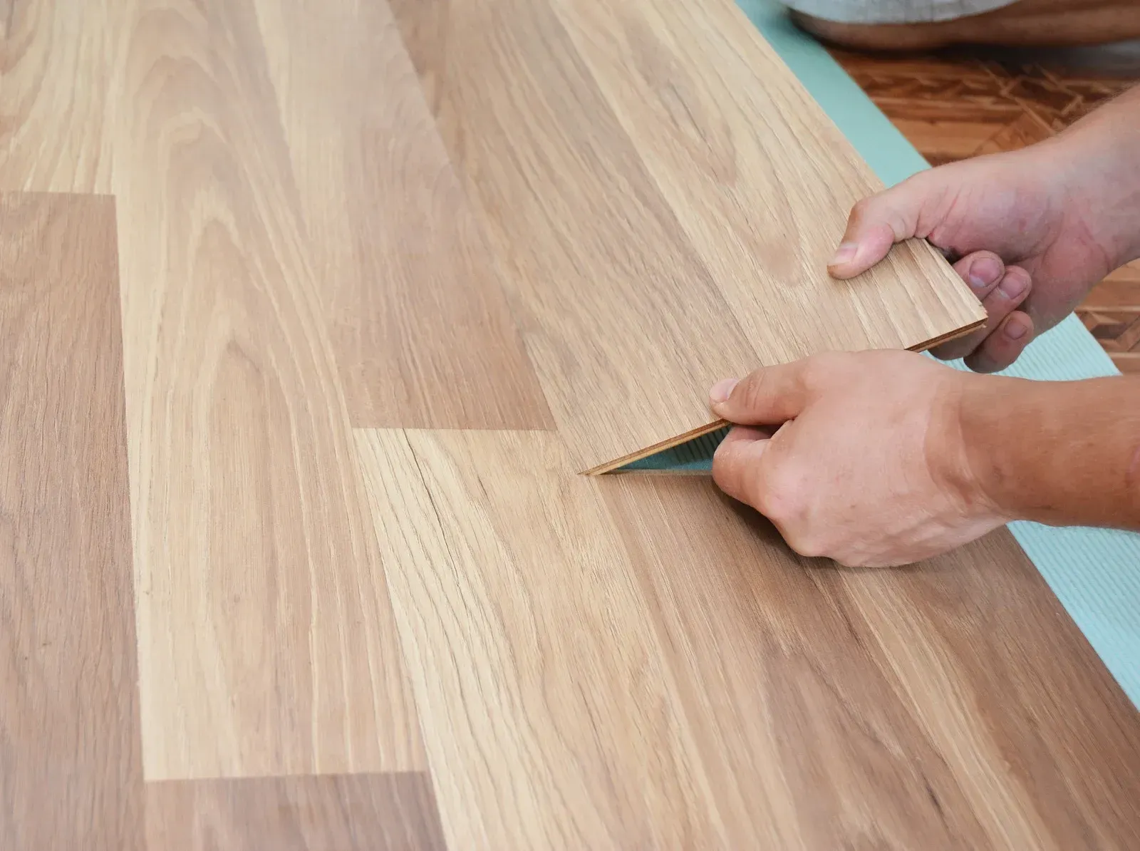Hands installing light wood-look laminate flooring planks over a pale green underlayment.
