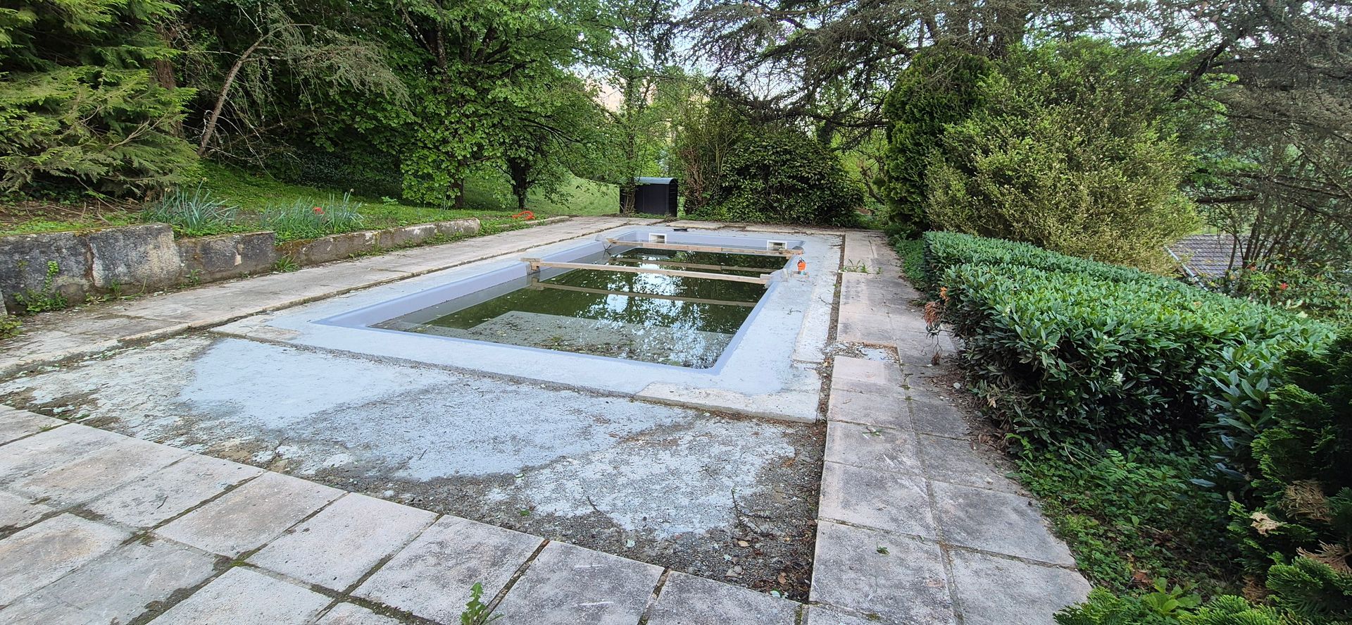 Un bassin rectangulaire aux eaux troubles, entouré de dalles de pierre et de verdure.