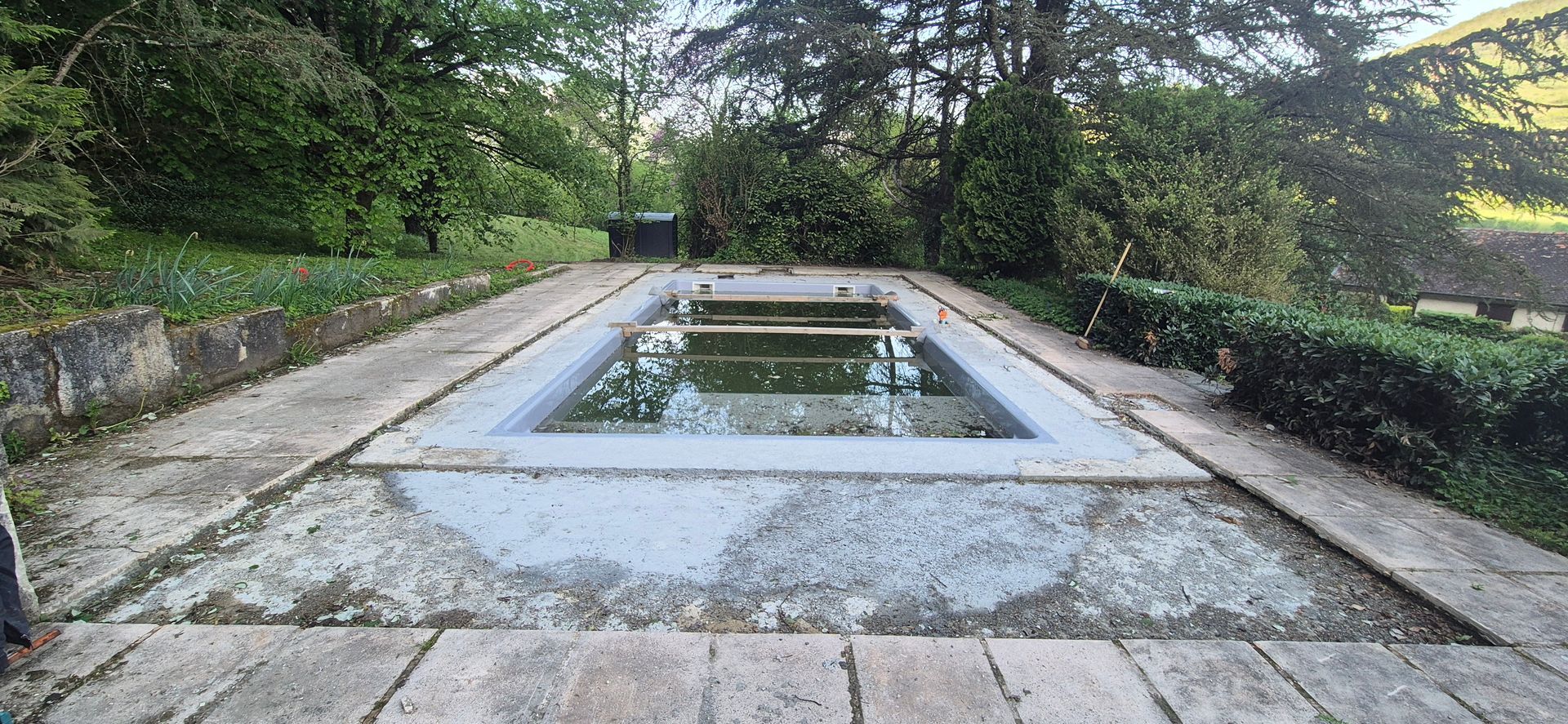 Piscine rectangulaire envahie par la végétation, entourée d'allées de pierre et d'une végétation luxuriante.