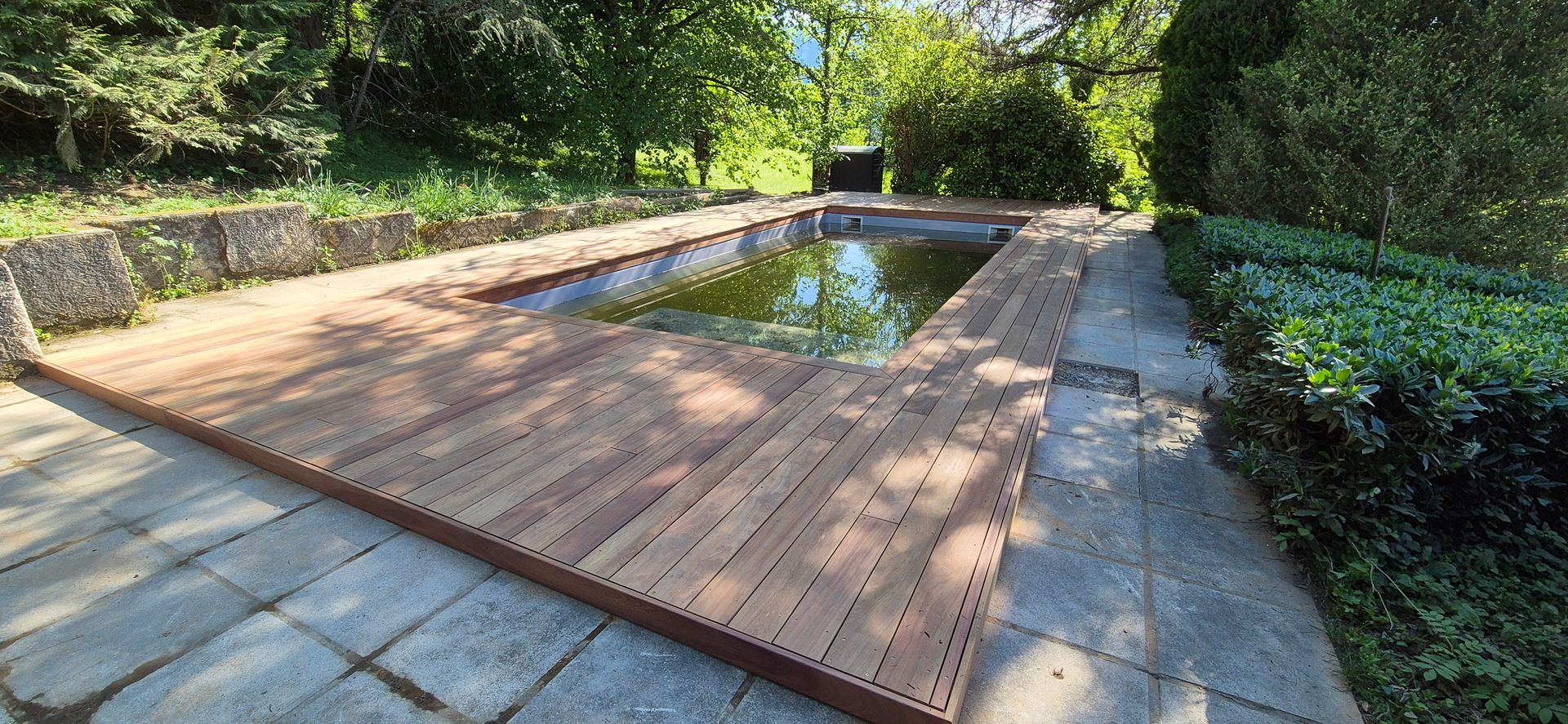Terrasse en bois entourant un bassin rectangulaire aux eaux vertes, le tout situé dans un jardin arboré.