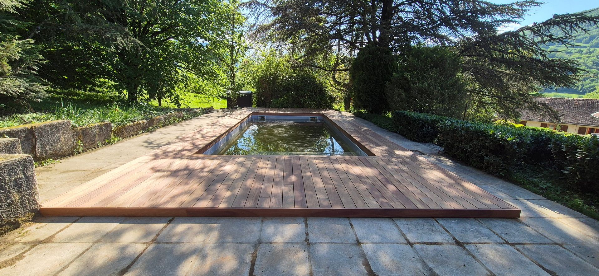 Une piscine extérieure rectangulaire avec terrasse en bois, entourée de verdure et de sentiers en pierre.