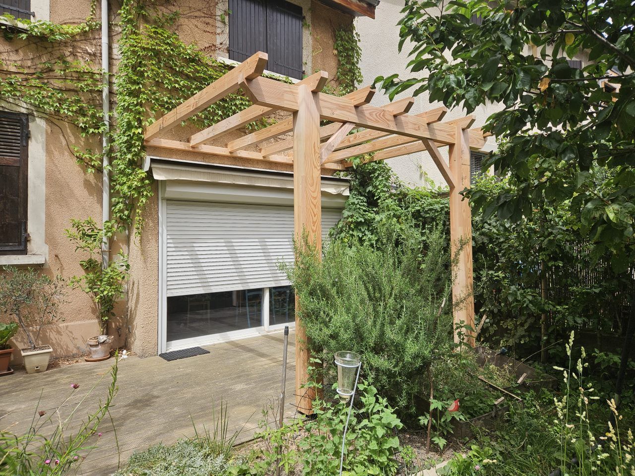Pergola en bois au-dessus d'une fenêtre munie d'un volet roulant blanc fermé, à côté d'un bâtiment orné de plantes grimpantes.