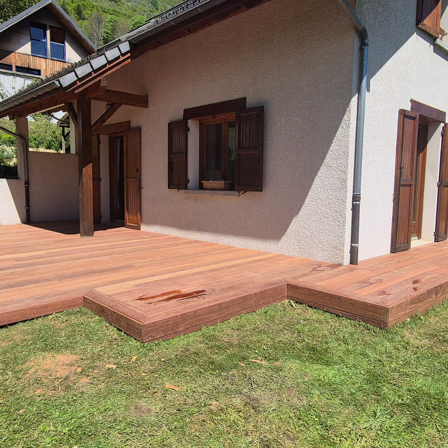 Terrasse en bois prolongeant une maison blanche aux volets et boiseries marron, située sur une pelouse verte.