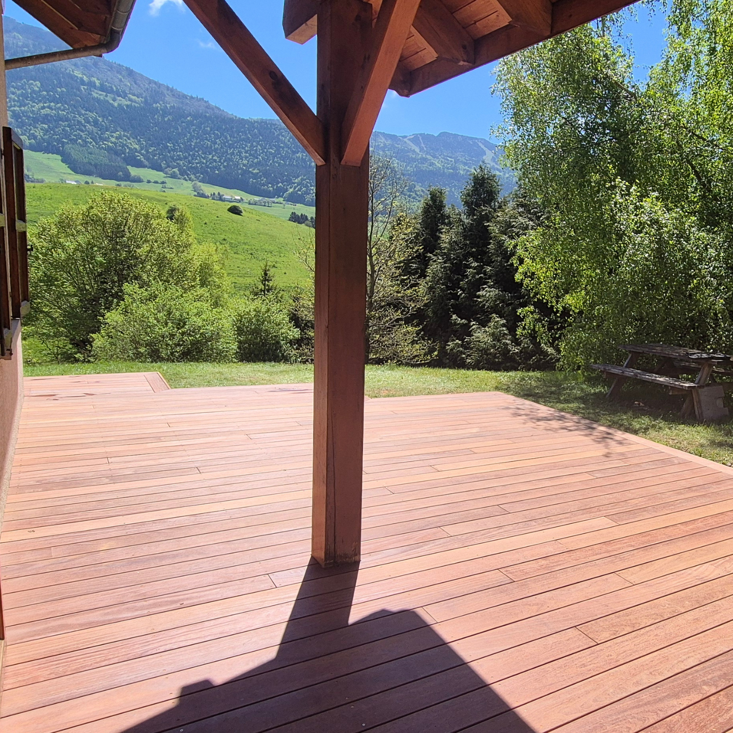 Terrasse en bois avec vue sur la montagne, abritée par un auvent en bois. Journée ensoleillée.