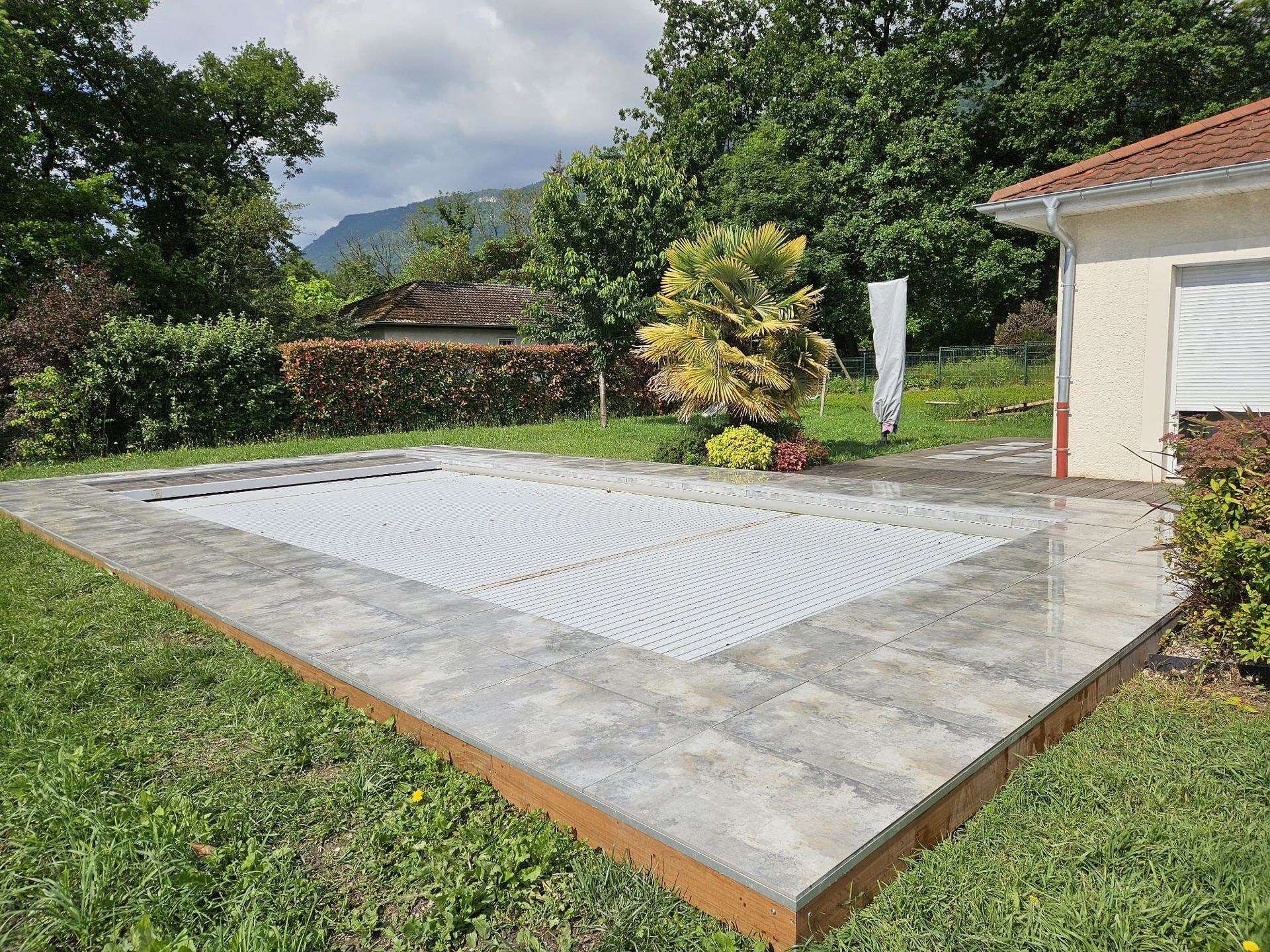 Une piscine rectangulaire recouverte d'un matériau gris, entourée de dalles de pierre, de pelouse et d'une maison.