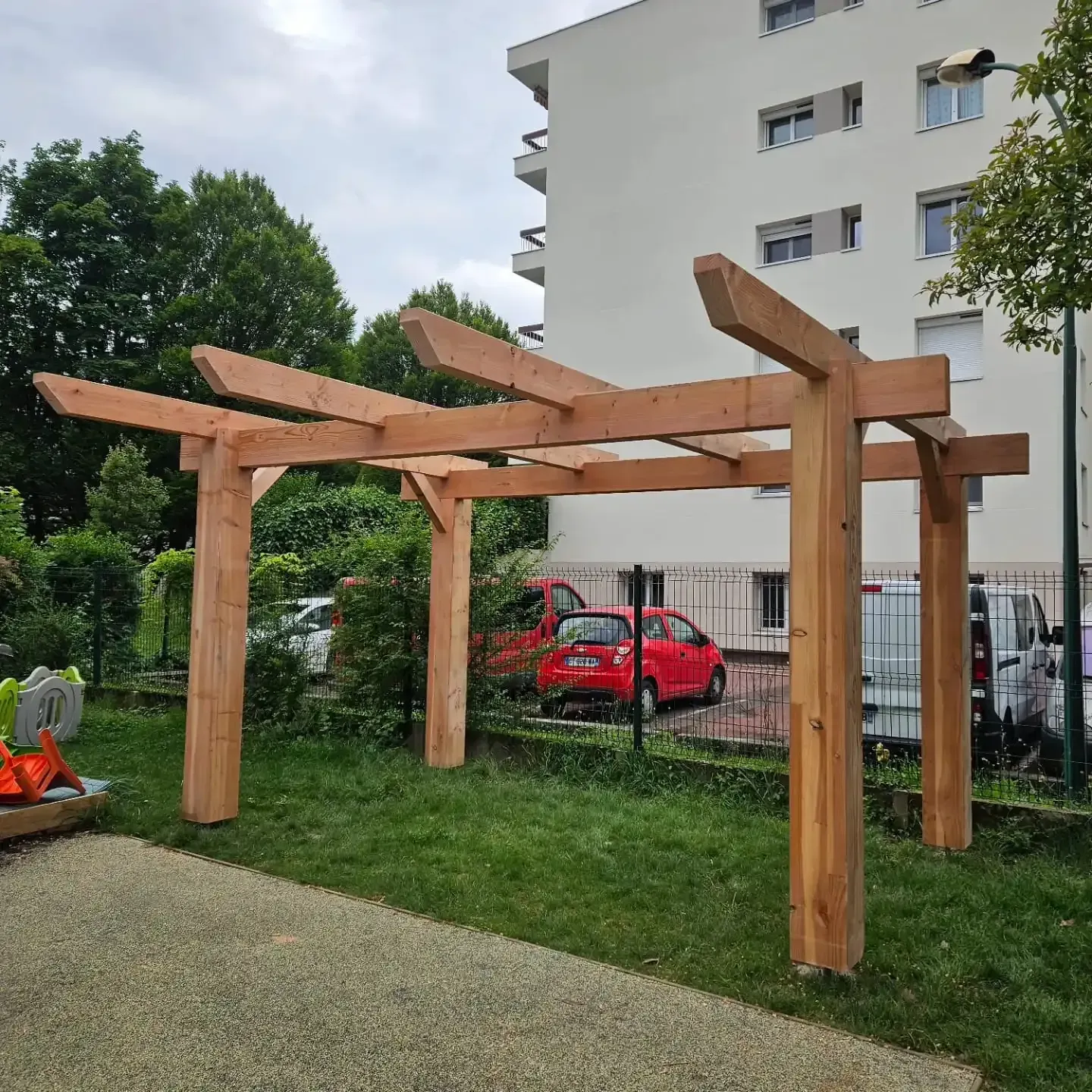 Une pergola en bois se dresse dans une zone herbeuse, avec un bâtiment en arrière-plan et une voiture rouge.