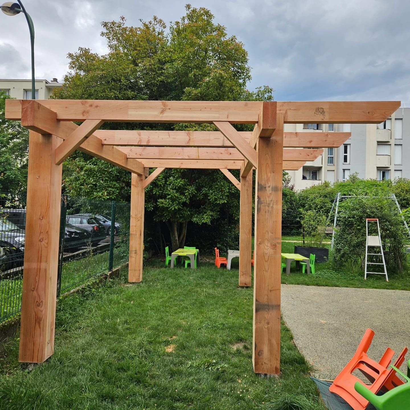 Pergola en bois dans un espace gazonné, avec de petites tables et des chaises visibles.