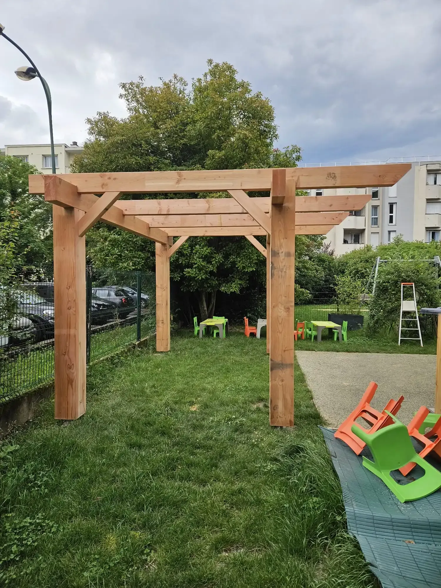 Pergola en bois dans une cour gazonnée, avec de petites tables et des chaises visibles. Ciel nuageux, cadre urbain.