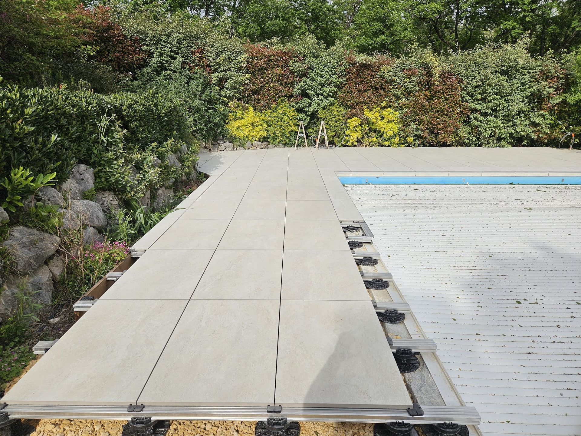 Terrasse en composite construite autour d'une piscine, entourée de verdure et d'un mur de pierre.