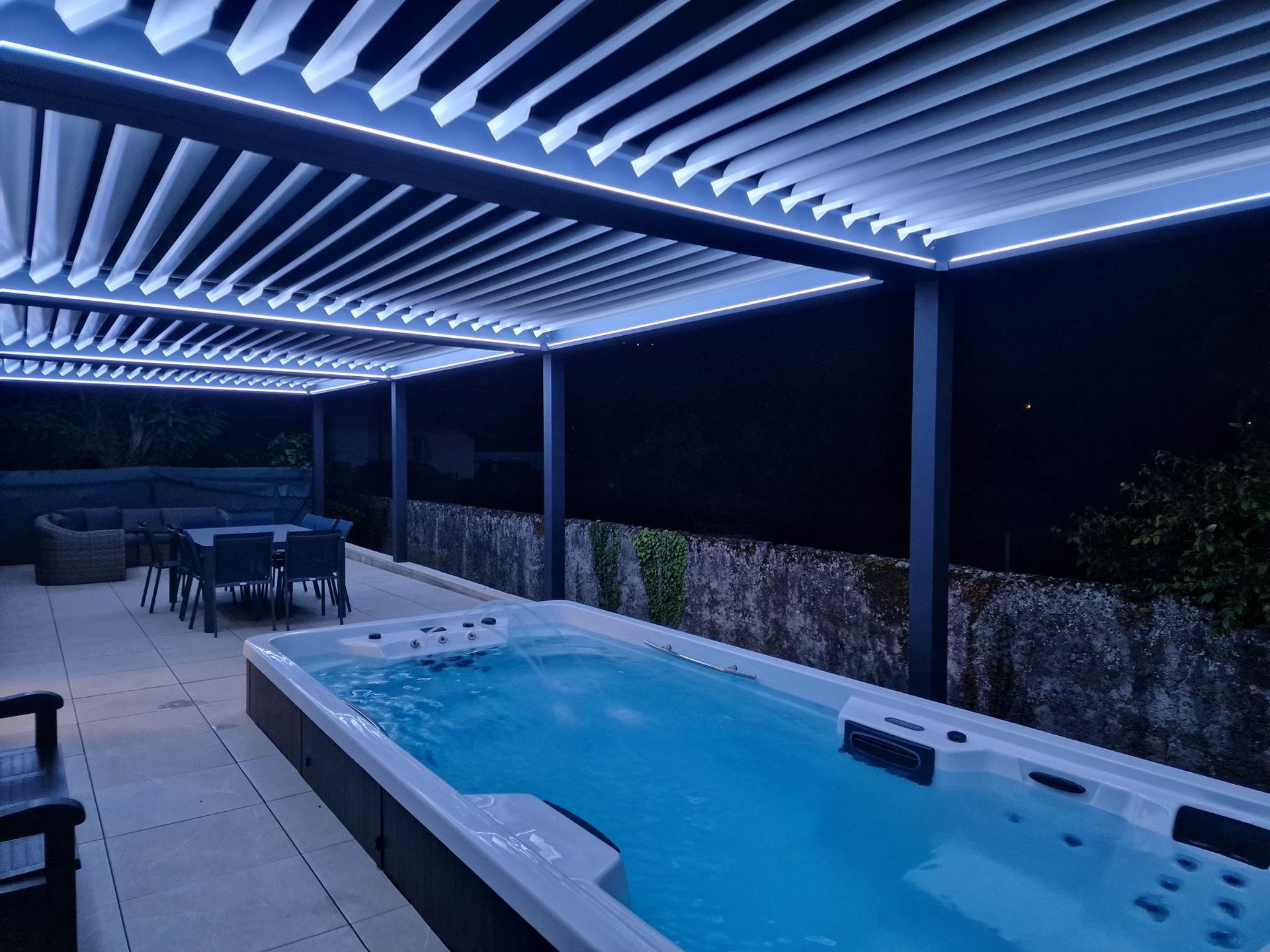 Patio avec pergola illuminée, spa de nage et coin salon le soir.