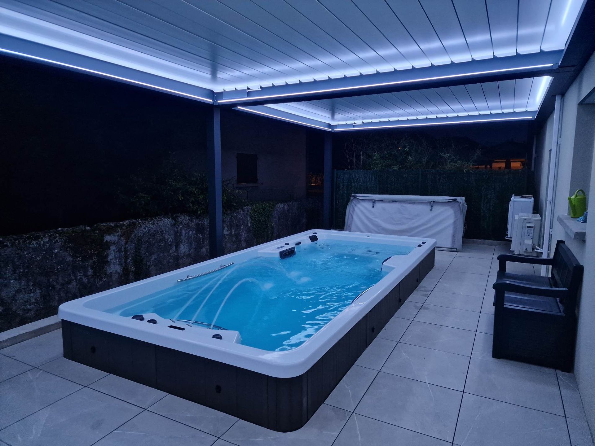 Piscine extérieure sous une pergola éclairée la nuit. Piscine brun foncé, carrelage blanc, mobilier noir.