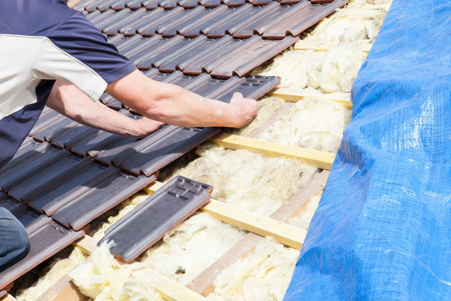 Worker installing roof insulation under dark roof tiles, with blue tarp at right.