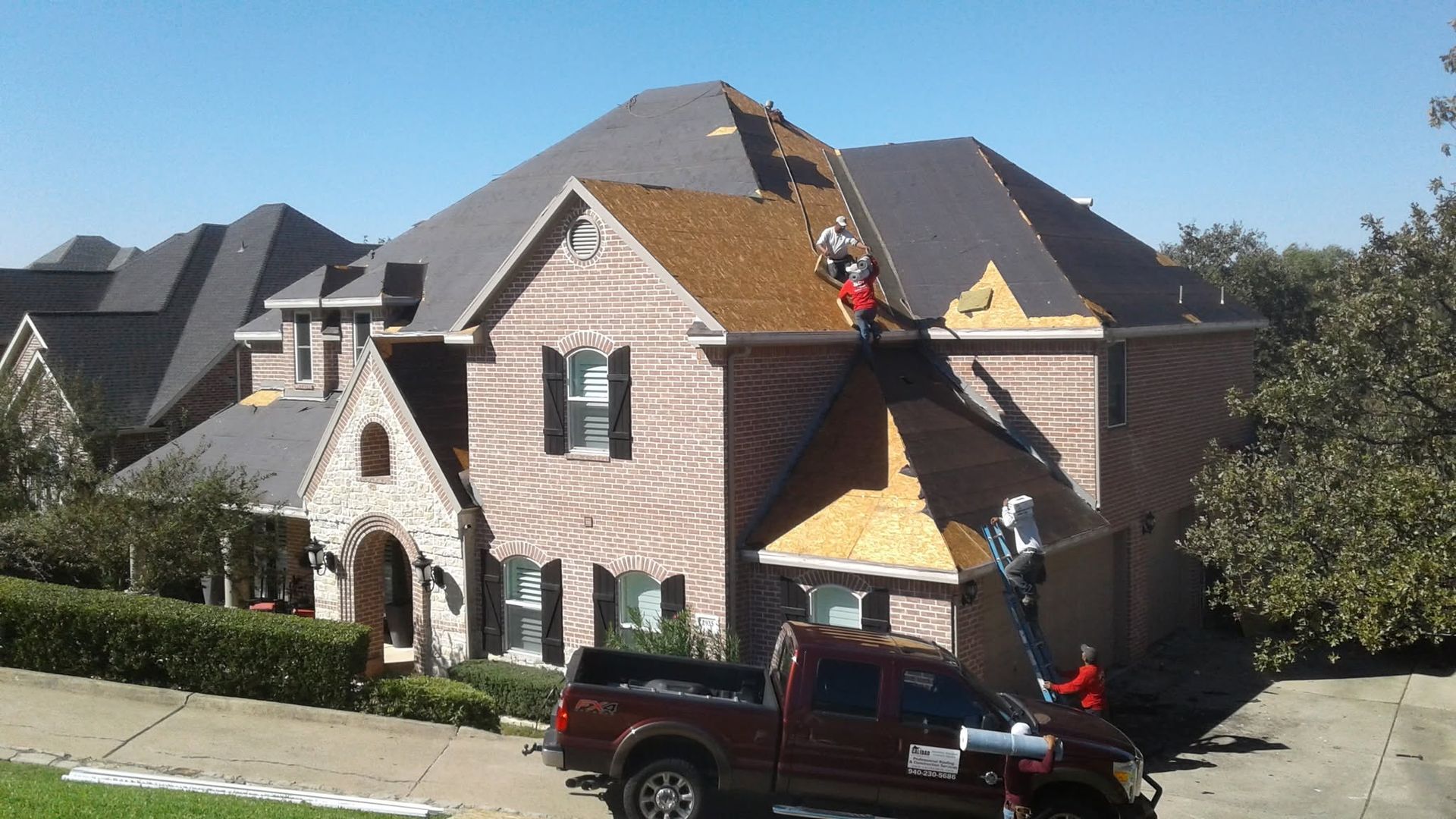 Workers repairing a tan house roof with a truck parked in the driveway