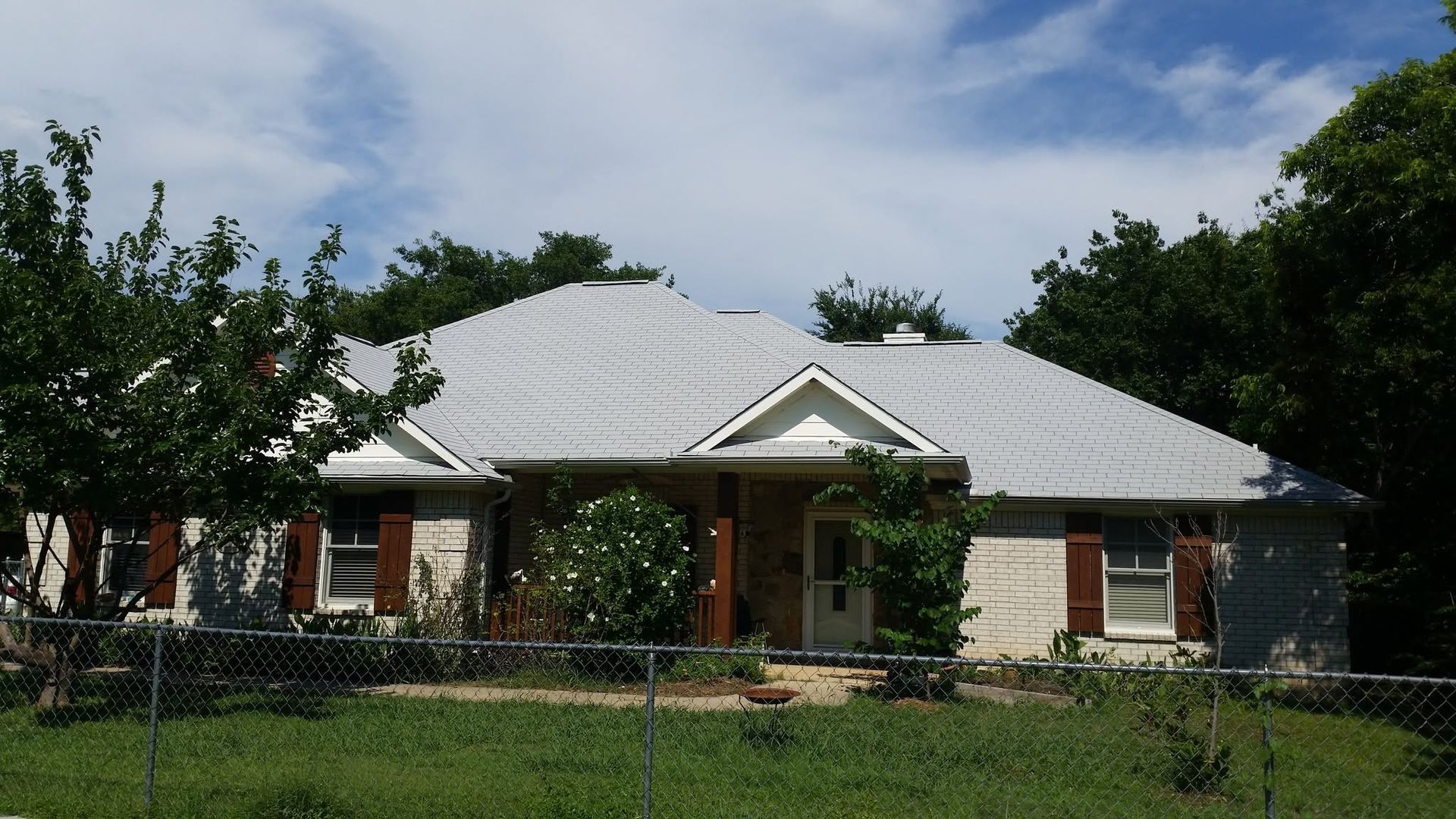 Single-story house with white roof, brick accents, and a front porch behind a fenced yard.
