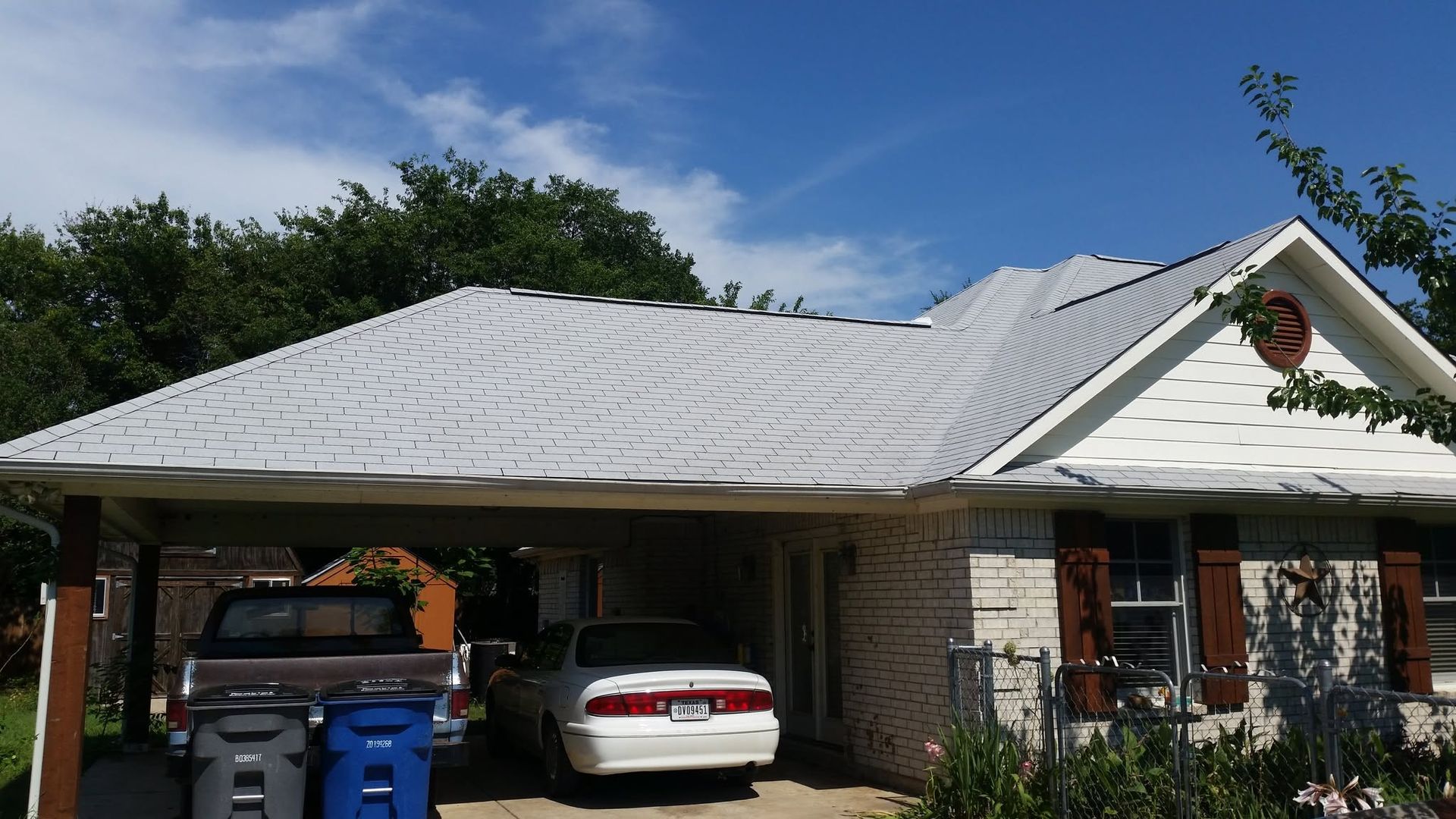 Single-story brick house with a white metal roof, parked cars, and blue trash bins in the driveway