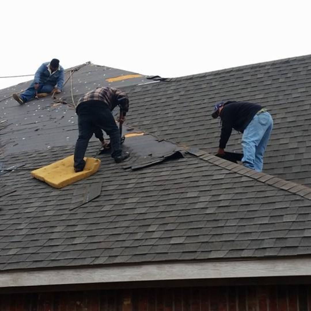 Three workers repairing a shingled roof, with tools and materials spread across the slope.