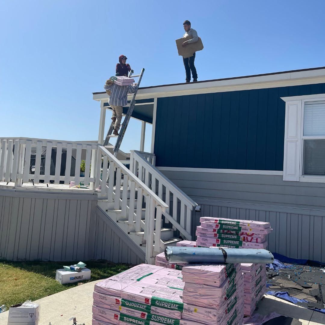 Workers on a rooftop near a ladder, with stacks of roofing shingles in the foreground.
