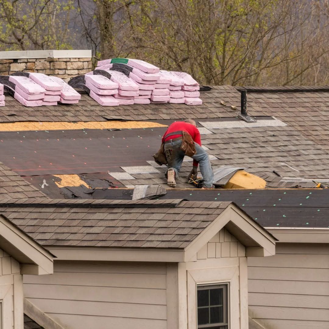 Roofing worker kneels on a shingled roof, repairing tiles near stacks of pink insulation.