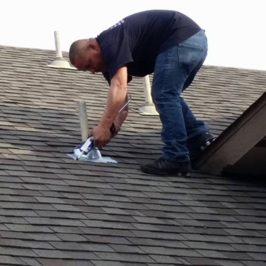 Worker crouched on a shingled roof, using a tool to repair the roof near a vent.