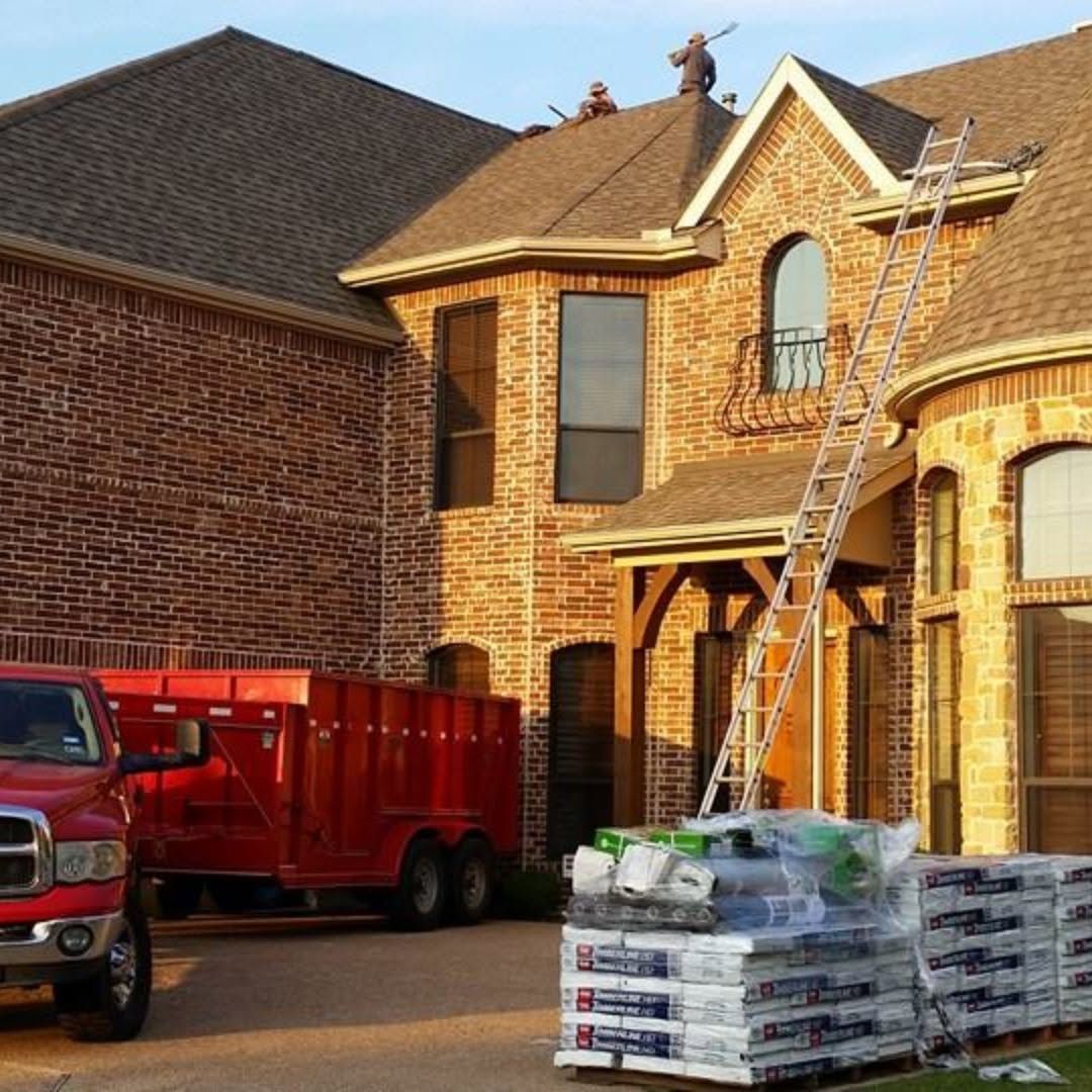 Brick house under construction with ladders, a red truck, and stacked roofing materials in the driveway