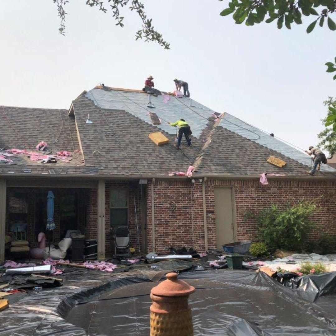 Workers repair a shingled roof on a brick house after storm damage, with debris in the yard.