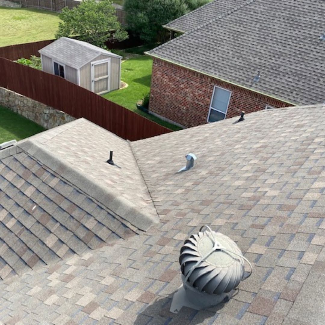 View from a rooftop over shingled roofs, a fenced backyard, and a small shed with trees beyond.