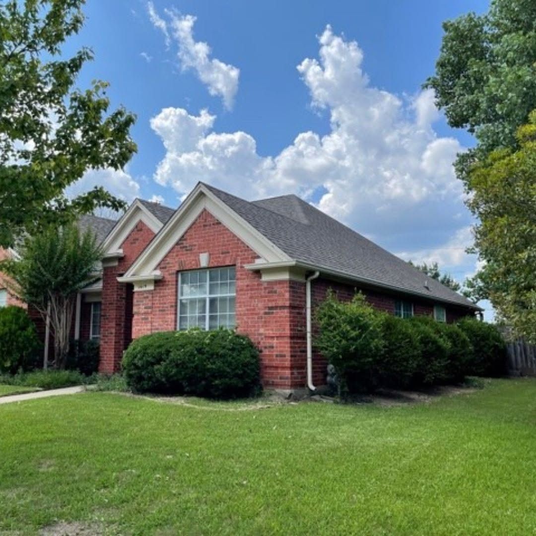 Red brick suburban house with gray roof, green lawn, and trees under a blue sky with clouds