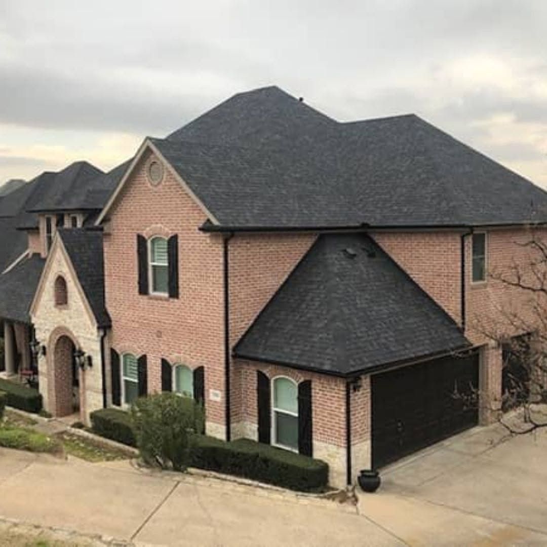 Large brick suburban house with dark shingled roof and attached garage under cloudy skies