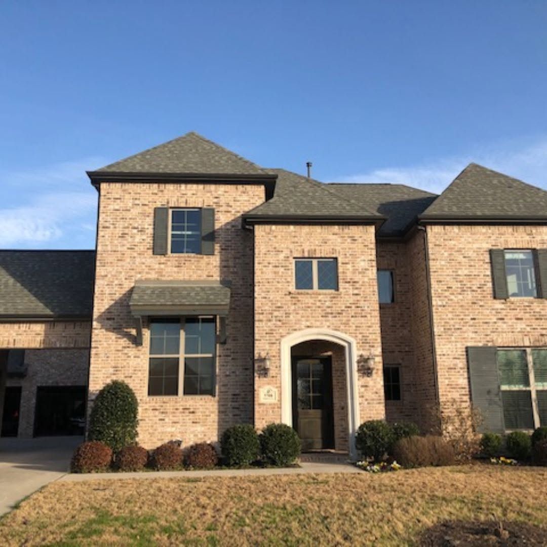 Two-story brick house with arched front door, dark shutters, and a front lawn under a blue sky